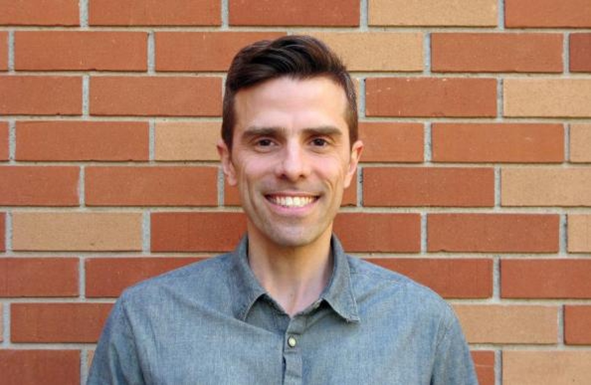 A man with dark brown eyes and dark brown hair wearing a blue button-up shirt stands in front of a brick wall and smiles.